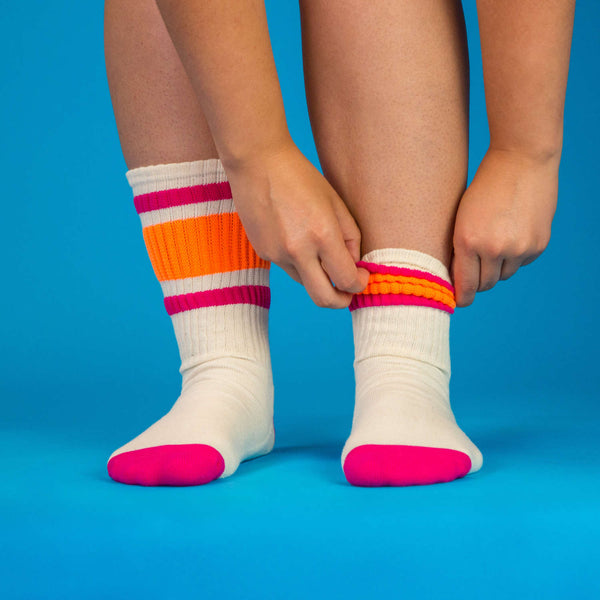 Lifestyle shot: Person putting on a pair of off-white crew socks with pink and orange stripes against a blue background. [lf]