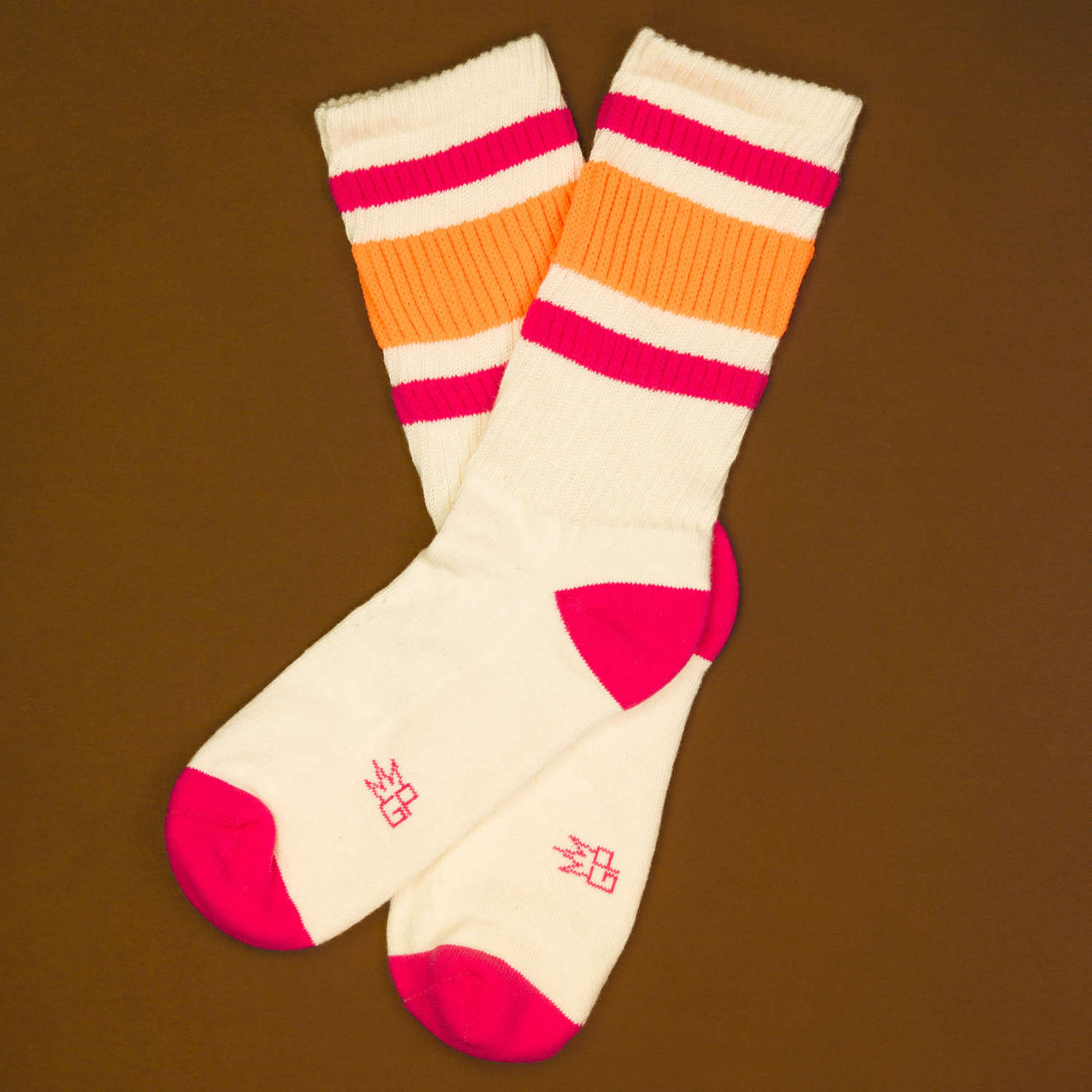 Flat lay: Pair of off-white crew socks with pink and orange stripes on a brown background. Socks are crossed. [lf]