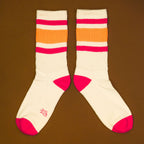 Flat lay: Pair of off-white crew socks with pink and orange stripes on a brown background. Socks are laid parallel. [lf]