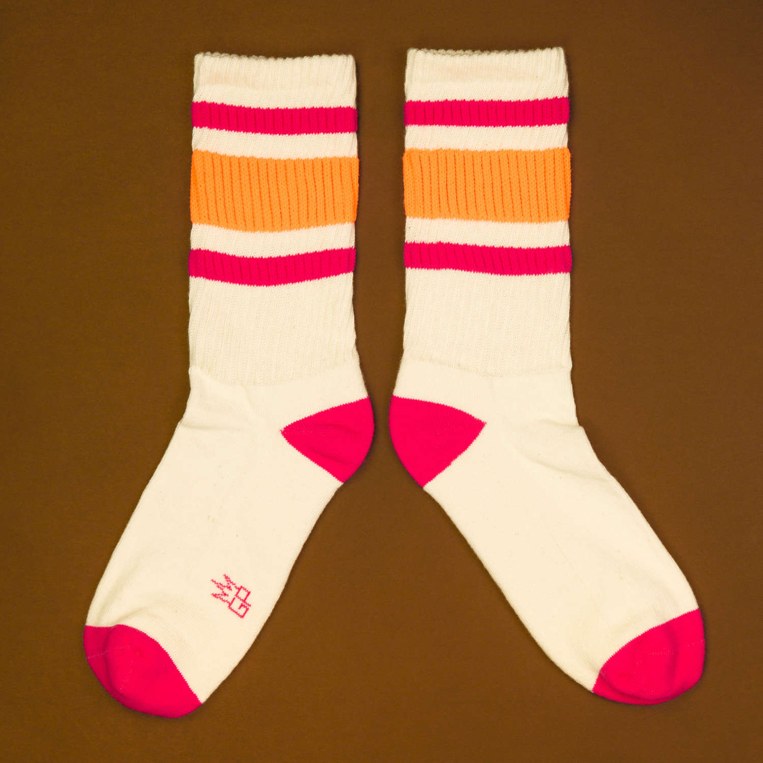 Flat lay: Pair of off-white crew socks with pink and orange stripes on a brown background. Socks are laid parallel. [lf]