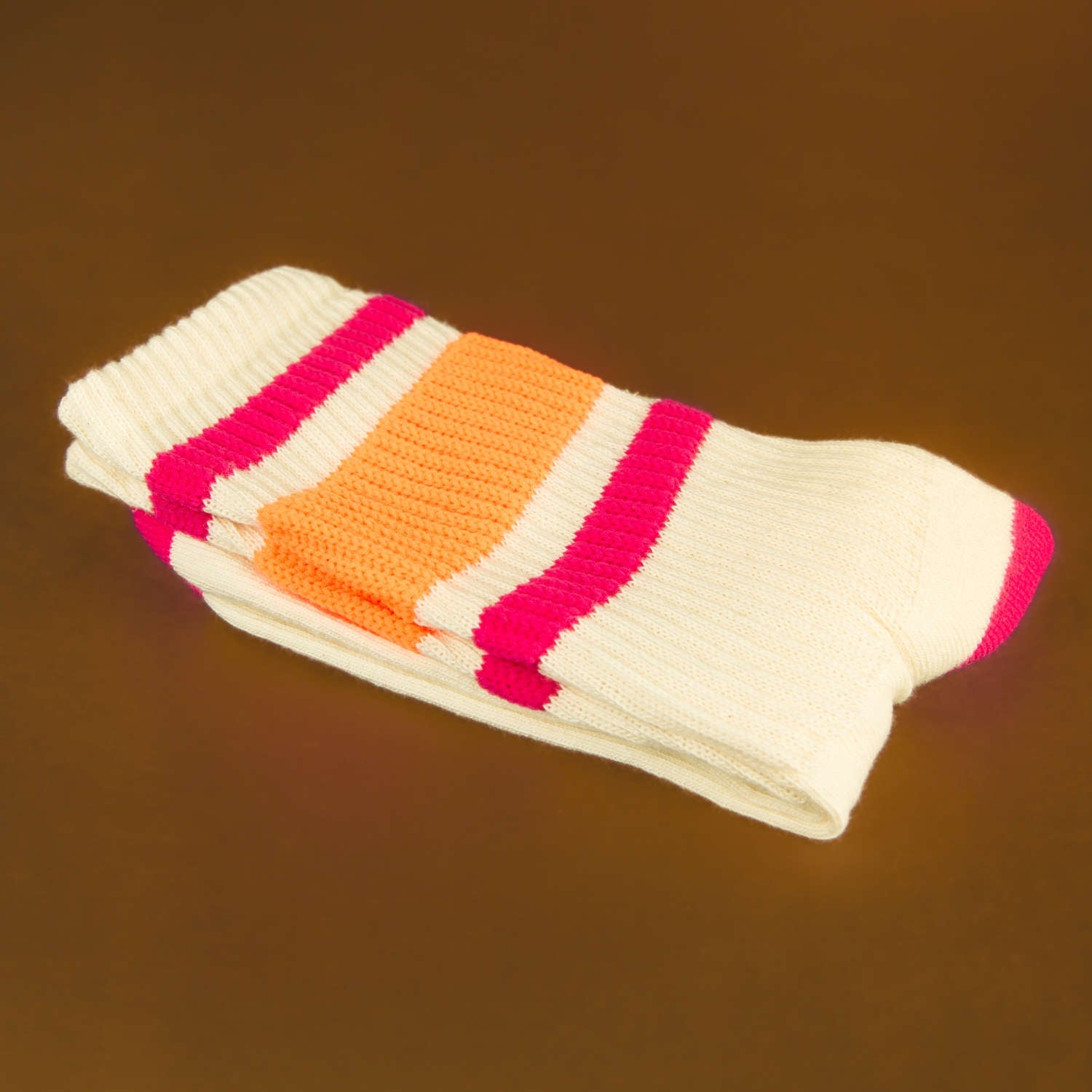 Close-up: Folded pair of off-white crew socks with pink and orange stripes on a brown background. [lf]
