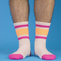 Lifestyle shot: Pair of off-white crew socks with pink and orange stripes are folded on a brown surface. [lf]