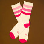 Flatlay product shot: Pair of cream crew socks with pink and red stripes on a brown background. [lf]