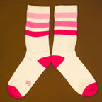 Flatlay product shot: Pair of cream crew socks with pink and red stripes laid out on a brown background. [lf]