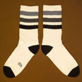 Flat lay product shot of a pair of off-white crew socks with black and grey stripes laid out on a brown background, angled. [lf]
