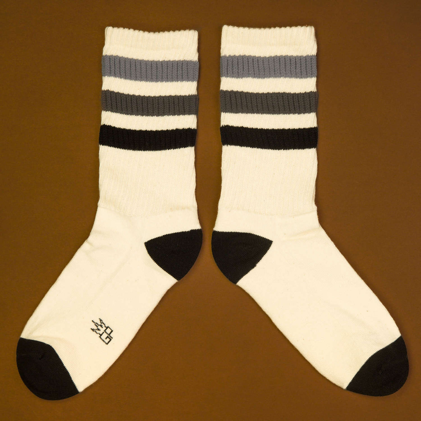 Flat lay product shot of a pair of off-white crew socks with black and grey stripes laid out on a brown background, angled. [lf]