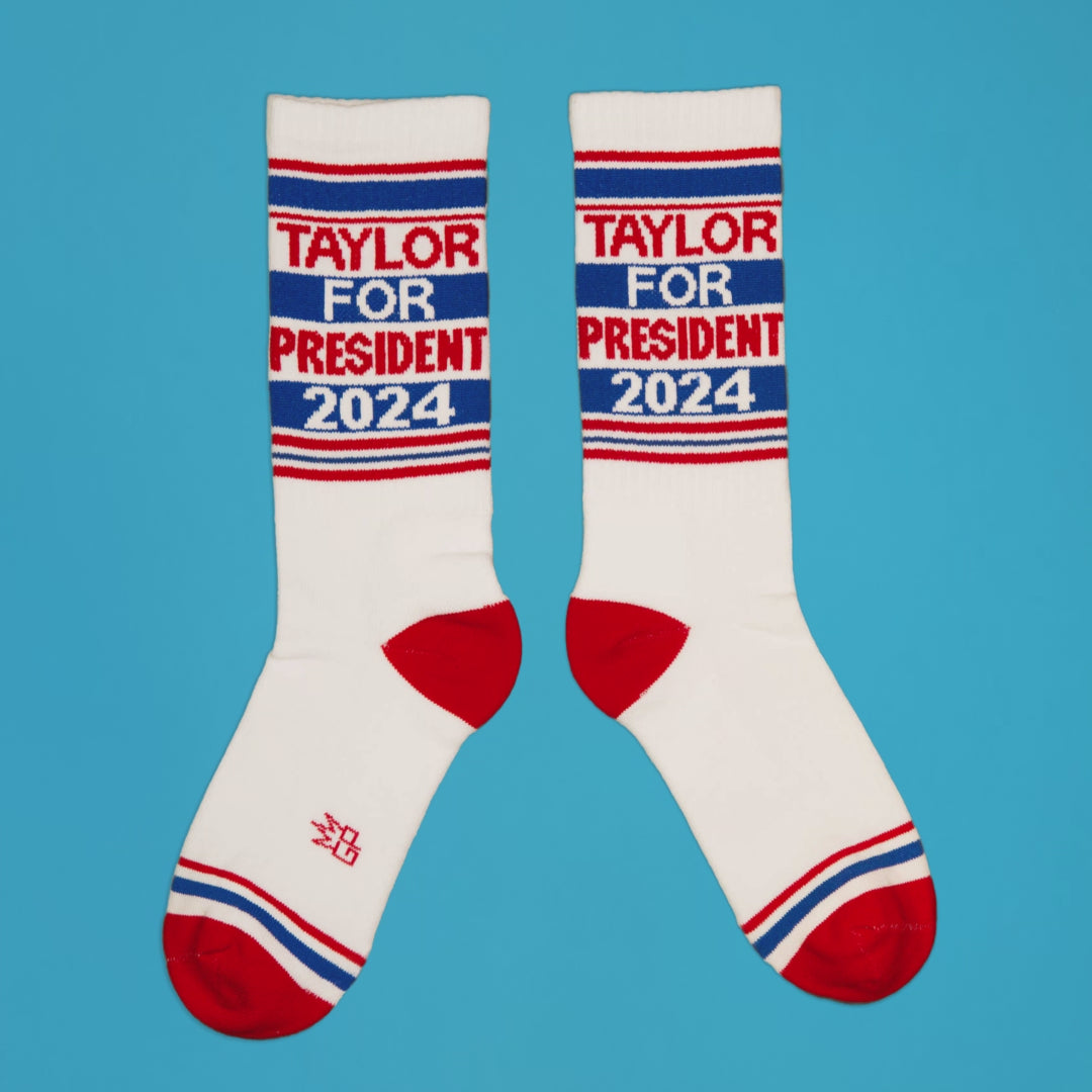Pair of 'Taylor for President 2024' socks displayed from a slightly elevated angle on a blue background. [lf]