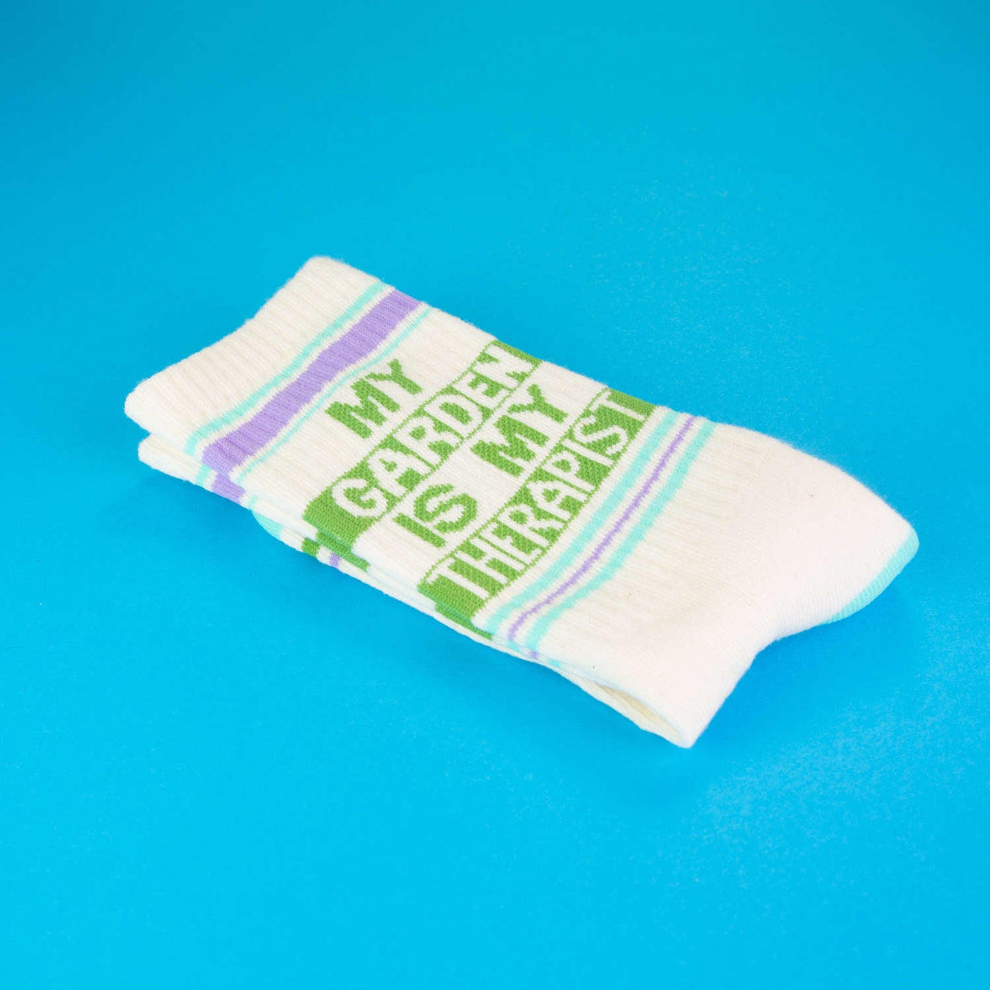 Close-up detail shot of 'My Garden Is My Therapist' socks folded on a bright blue background. [lf]