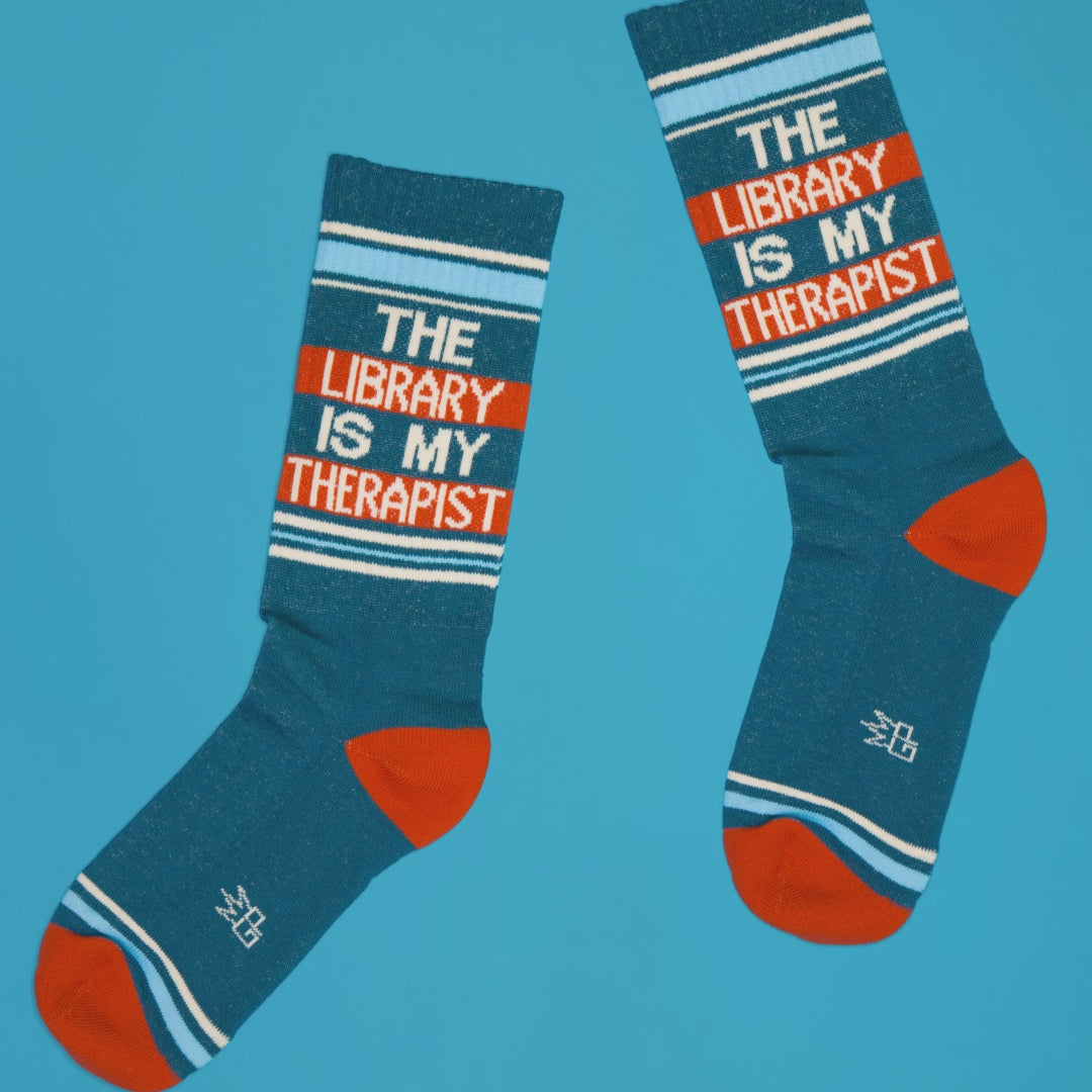 Pair of teal 'The Library Is My Therapist' socks on a blue background, angled view. [lf]