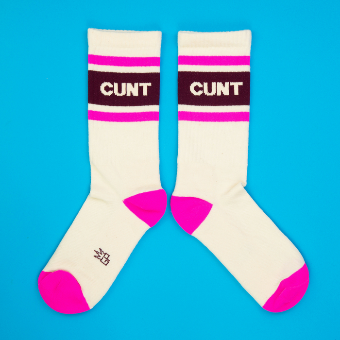 Flatlay of two off-white crew socks with pink accents and a dark maroon word on a bright blue background.