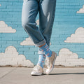 Person wearing light blue flying pig crew socks with cuffed jeans and beige sneakers by a cloud mural. [lf]
