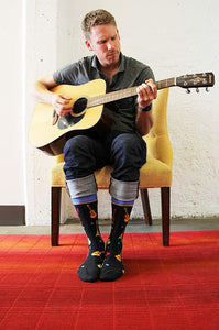 A man is sitting on a yellow chair playing an acoustic guitar. He is wearing a gray shirt, blue jeans, and colorful socks with a guitar pattern. He is sitting on a red rug with his feet crossed.