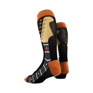 A person wearing black jeans and dress shoes is sitting on the ground with an acoustic guitar resting on their right leg. The person is wearing a pair of socks that are black with an orange musical staff and brown fretboard design.