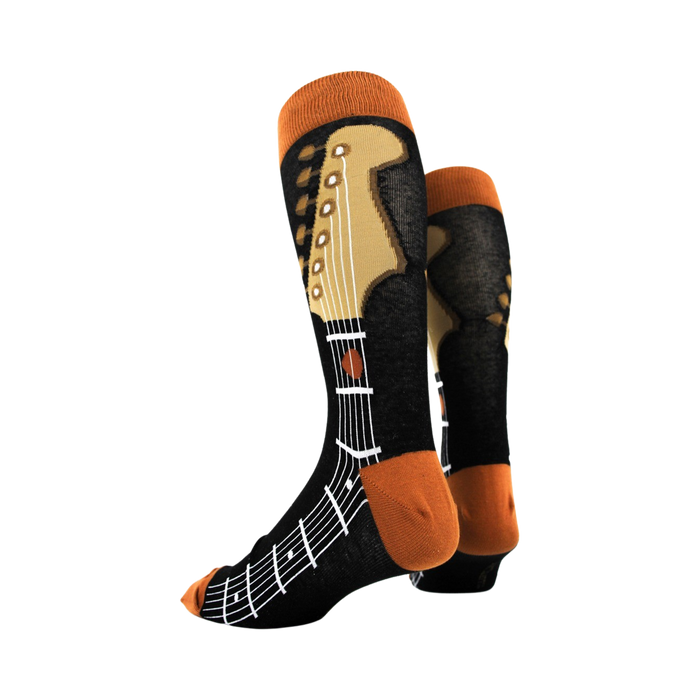 A person wearing black jeans and dress shoes is sitting on the ground with an acoustic guitar resting on their right leg. The person is wearing a pair of socks that are black with an orange musical staff and brown fretboard design.