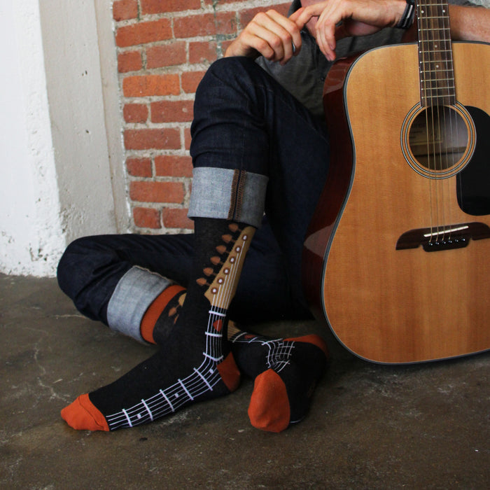 A person wearing black jeans and dress shoes is sitting on the ground with an acoustic guitar resting on their right leg. The person is wearing a pair of socks that are black with an orange musical staff and brown fretboard design.