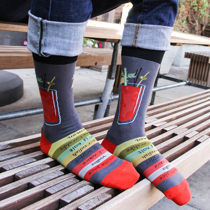 A person is sitting on a wooden bench with their feet resting on the seat. They are wearing blue jeans and socks with a bloody mary cocktail design. The socks are gray with a red and green cocktail graphic and multi-colored striped toes and heels.