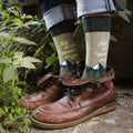 Lifestyle shot of Wanderer socks on a person's feet, paired with brown leather boots, outdoors near plants. [lf]