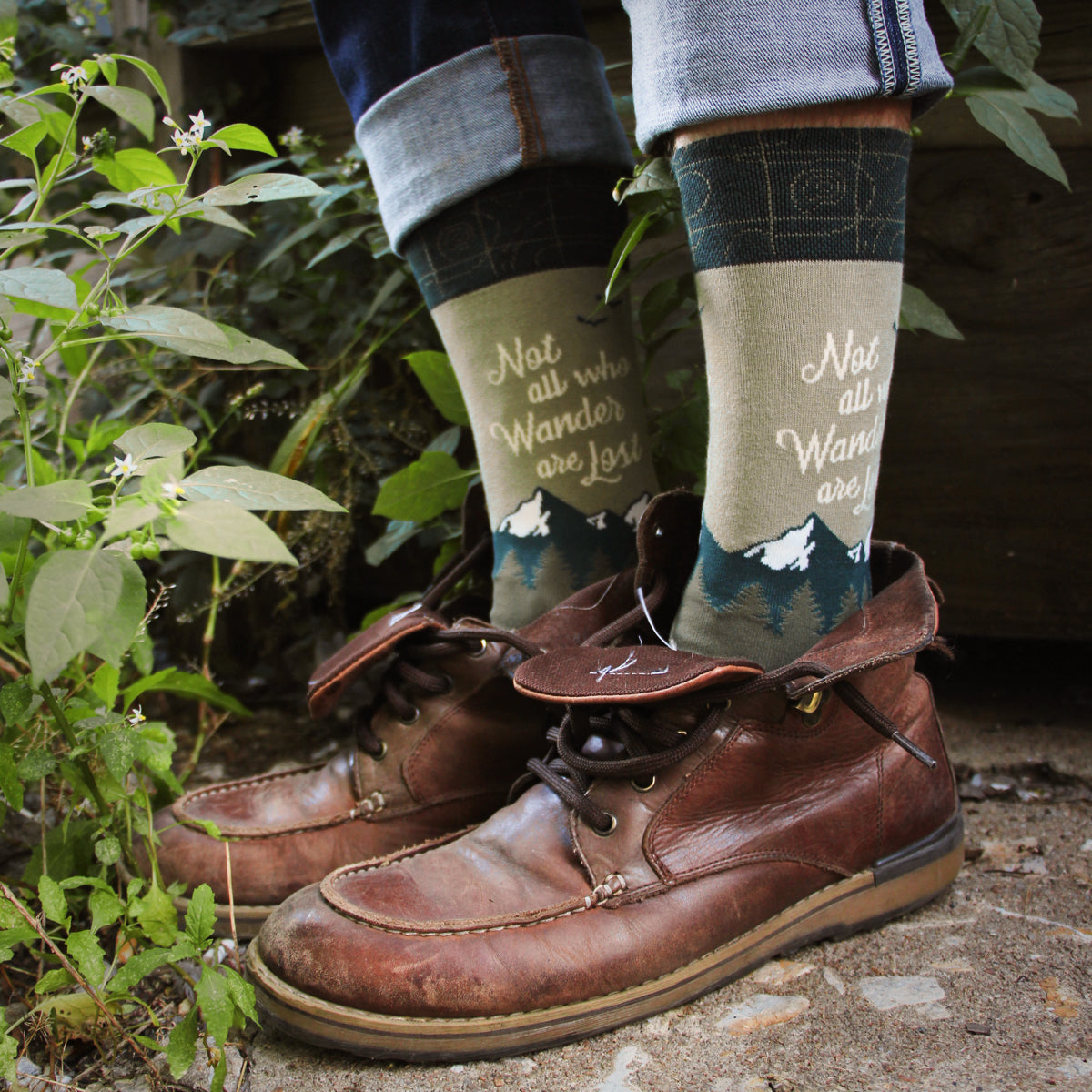Lifestyle shot of Wanderer socks on a person's feet, paired with brown leather boots, outdoors near plants. [lf]