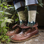 Lifestyle shot of Wanderer socks on a person's feet, paired with brown leather boots, outdoors near plants. [lf]