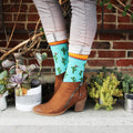 Lifestyle shot of Cactus socks worn with brown ankle boots. The socks are paired with succulents and a brick wall. [lf]