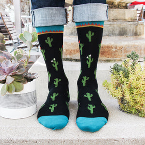 A person is modeling a pair of black socks with a pattern of green cacti wearing pink flowers. The socks have bright blue toes and heels. The person is wearing blue jeans and brown leather shoes. There are two planters with green succulent plants on the ground next to the person's feet.