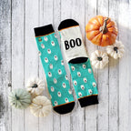 Flatlay product shot of teal ghost socks with a 'BOO' detail, styled with mini pumpkins on a white wooden background. [lf]
