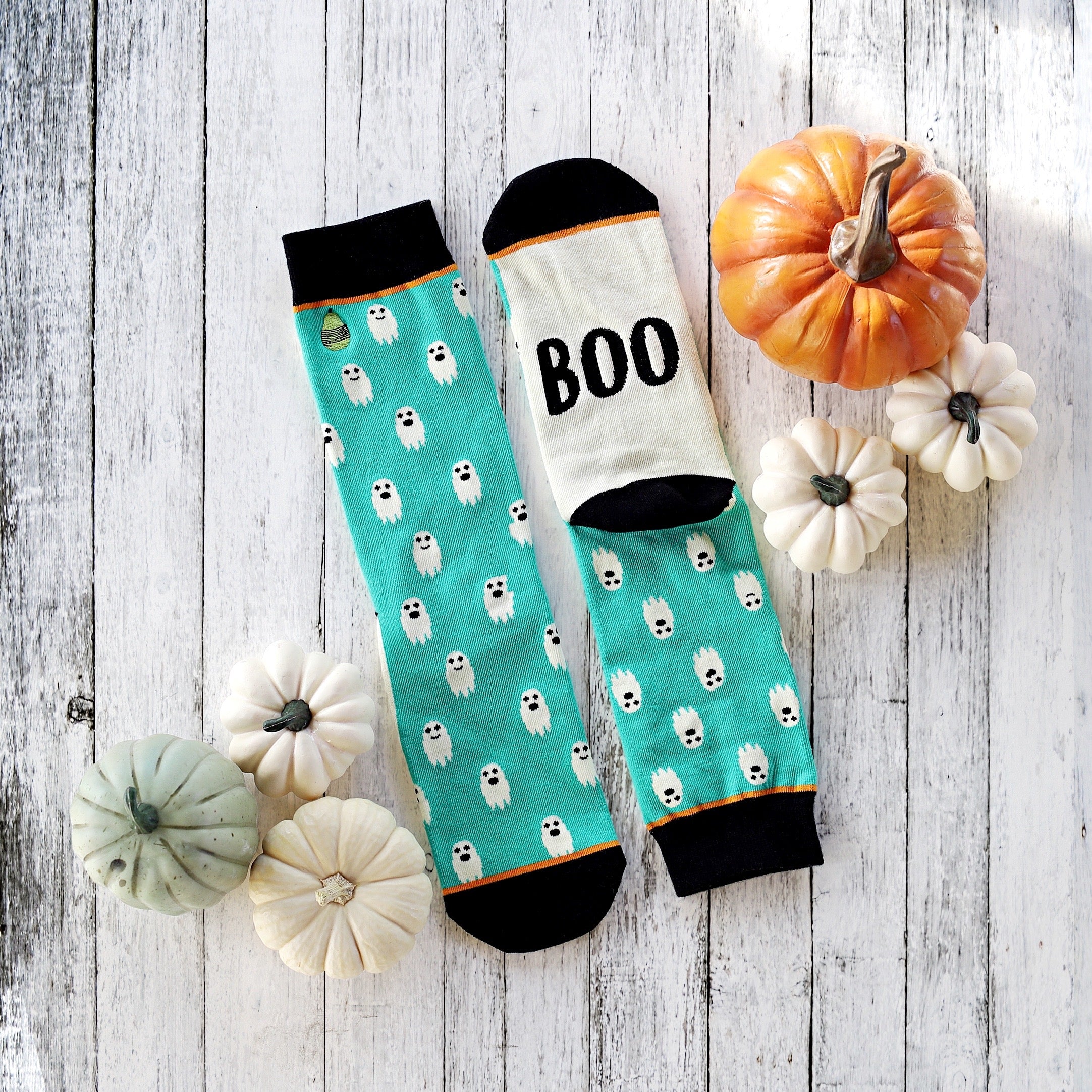 Flatlay product shot of teal ghost socks with a 'BOO' detail, styled with mini pumpkins on a white wooden background. [lf]