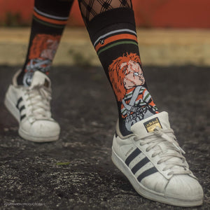 A person is shown from the waist down wearing dark blue jeans, black sneakers, and socks with a pattern of the horror movie character Chucky. The person is holding a knife.