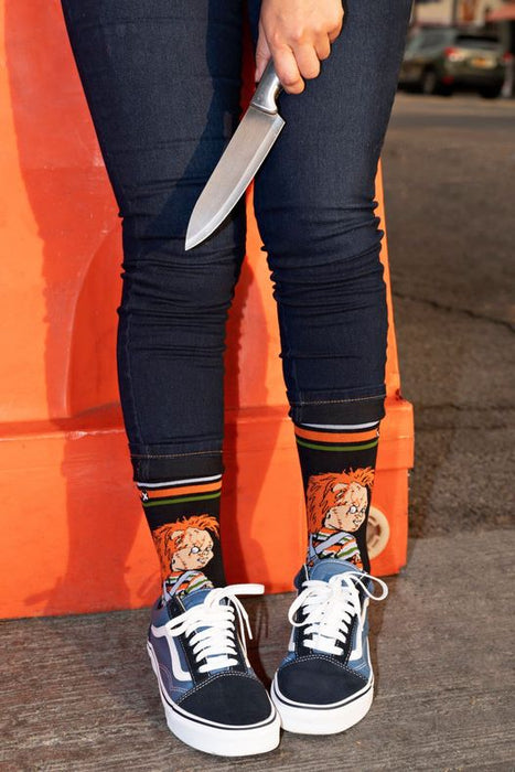 A person is shown from the waist down wearing dark blue jeans, black sneakers, and socks with a pattern of the horror movie character Chucky. The person is holding a knife.