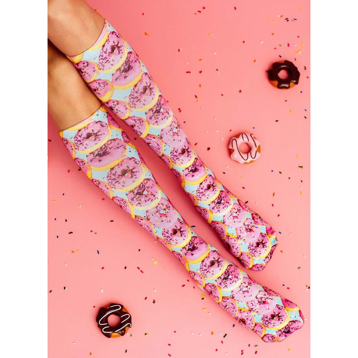 A pair of legs is shown from the knees down. The person is wearing socks with a pattern of pink icing topped donuts with rainbow sprinkles on a blue background. The socks are pulled up to just below the knees. The person is standing on a pink background with a few donut images and many sprinkles scattered on the surface.