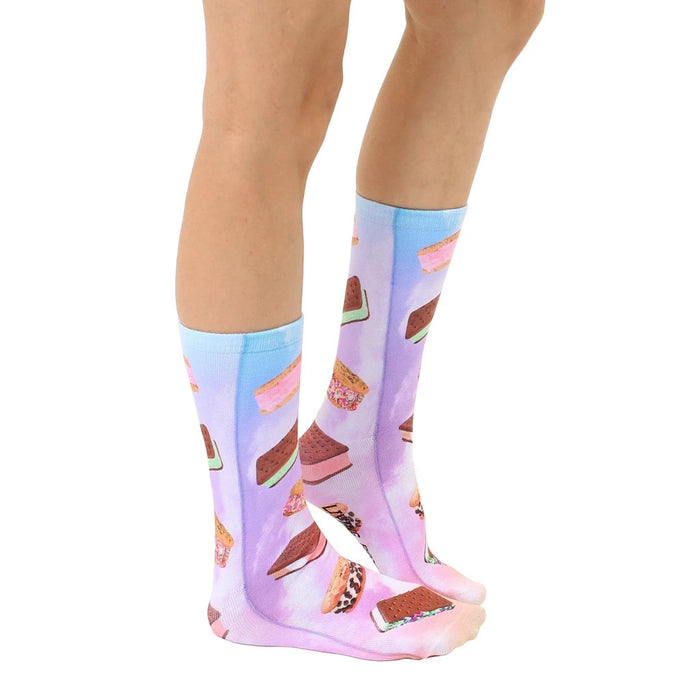 A pair of legs is shown from the knees down wearing a pair of knee-high socks with a light blue toe and heel and a pattern of various ice cream sandwiches against a pastel purple background.