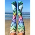 Lifestyle shot: Person wearing rainbow mermaid scale knee-high socks on a beach. Ocean and sky in the background. [lf]