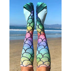 Lifestyle shot: Person wearing rainbow mermaid scale knee-high socks on a beach. Ocean and sky in the background. [lf]