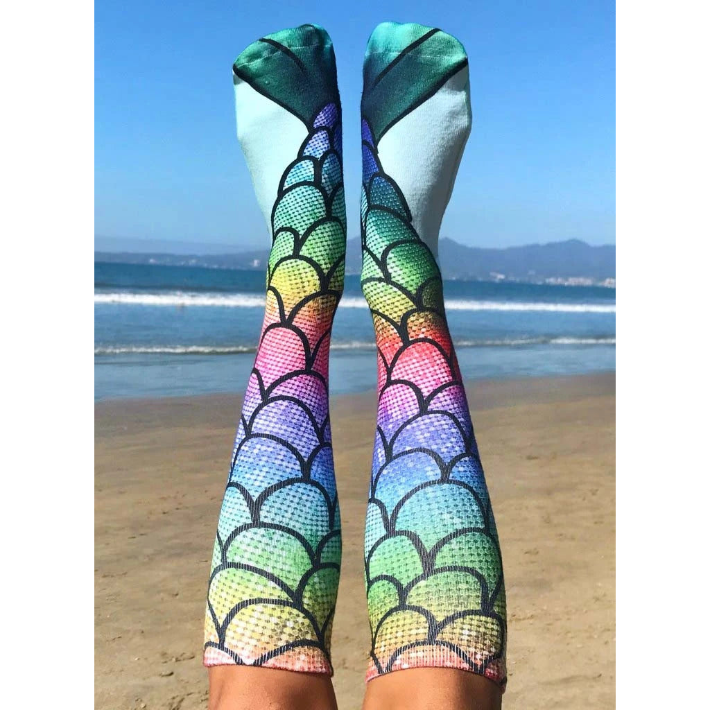 Lifestyle shot: Person wearing rainbow mermaid scale knee-high socks on a beach. Ocean and sky in the background. [lf]