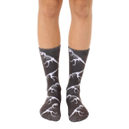 A pair of crew socks on a person's legs. The dark grey socks feature a repeated pattern of white T-Rex dinosaur skeletons. [lf]