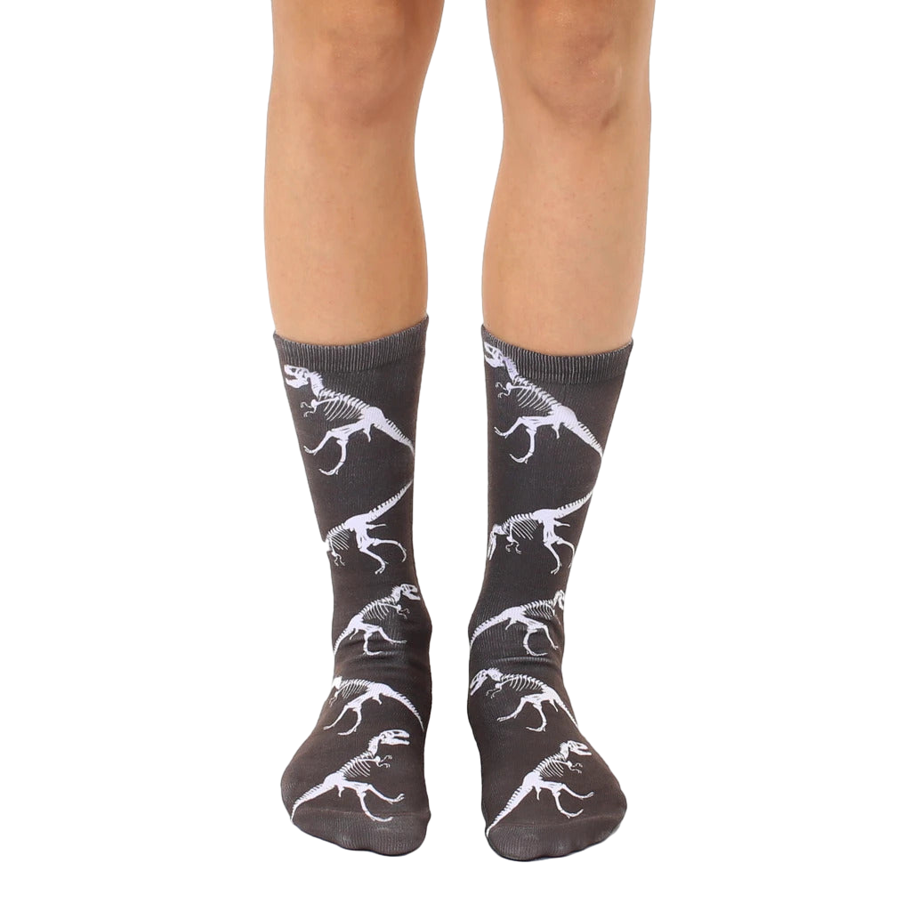 A pair of crew socks on a person's legs. The dark grey socks feature a repeated pattern of white T-Rex dinosaur skeletons. [lf]