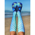 Lifestyle shot of Mermaid Blue socks worn by a person sitting on a beach, showing the socks in a relaxed, everyday setting. [lf]