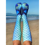 Lifestyle shot of Mermaid Blue socks worn by a person sitting on a beach, showing the socks in a relaxed, everyday setting. [lf]