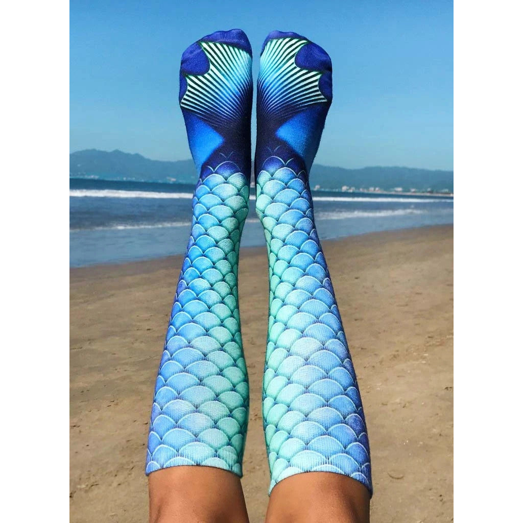 Lifestyle shot of Mermaid Blue socks worn by a person sitting on a beach, showing the socks in a relaxed, everyday setting. [lf]