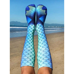 A pair of blue and green mermaid tail socks are shown from the knees down. The wearer is standing on a beach with the ocean and mountains in the background.