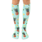 Pair of knee-high socks. Teal background with repeating pattern of cartoon pineapples with googly eyes and pineapple slices. [lf]