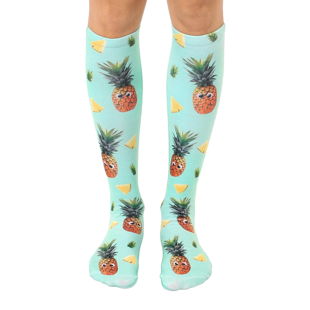 Pair of knee-high socks. Teal background with repeating pattern of cartoon pineapples with googly eyes and pineapple slices. [lf]