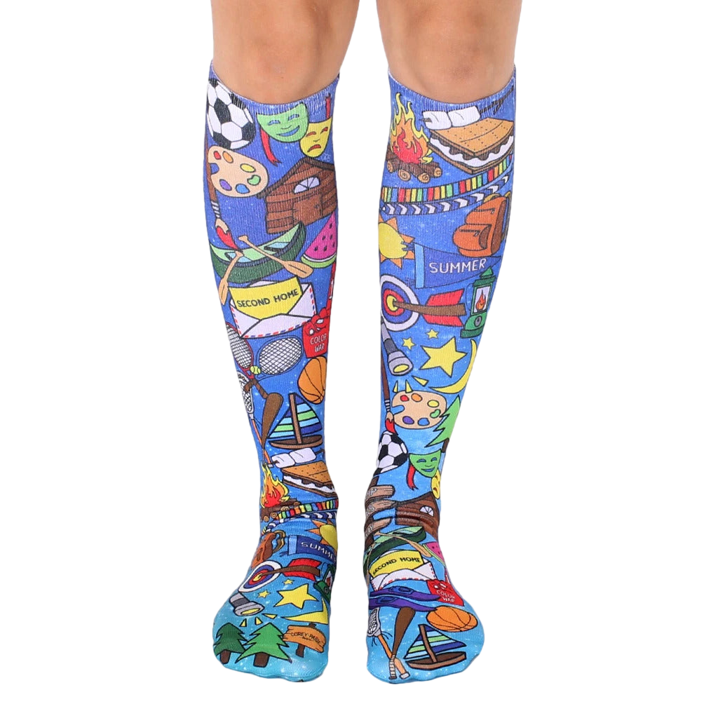 A pair of knee-high socks on a model's legs.  The socks are blue with colorful illustrations of summer camp activities, including sports equipment, campfires, and s'mores. [lf]