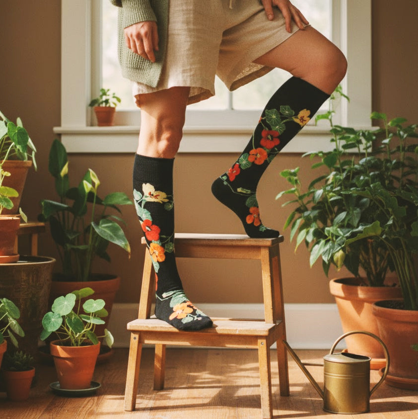 Black knee-high floral socks with red and yellow nasturtiums worn by a model in a sunlit room with plants. [lf]
