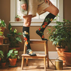 Black knee-high floral socks with red and yellow nasturtiums worn by a model in a sunlit room with plants. [lf]