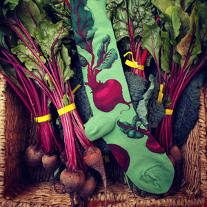 A green sock with a pattern of red beets on it is laying in a basket of red beets and beet greens.