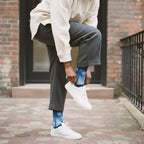 Model wearing blue gradient crew socks with white snowflakes, grey trousers, and white sneakers on a brick-paved sidewalk. [lf]