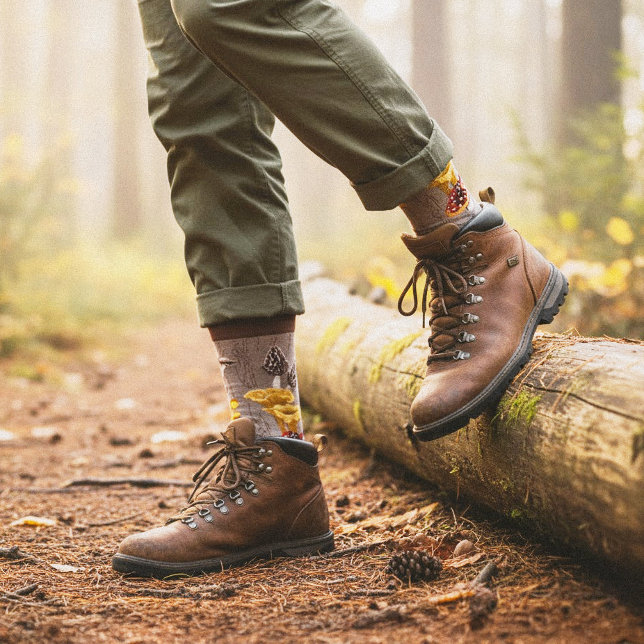 Model wears taupe mushroom crew sock, brown hiking boots, olive pants, stepping on a mossy log in a foggy forest. [lf]
