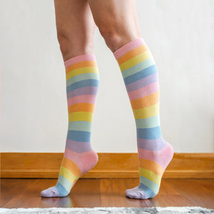A person's legs are shown from the knees down. The person is wearing pastel rainbow striped socks and standing on a brown wooden floor.