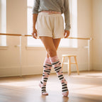 Person in white shorts, pink knee-high socks with black and white bandage stripes, standing in a ballet pose indoors. [lf]